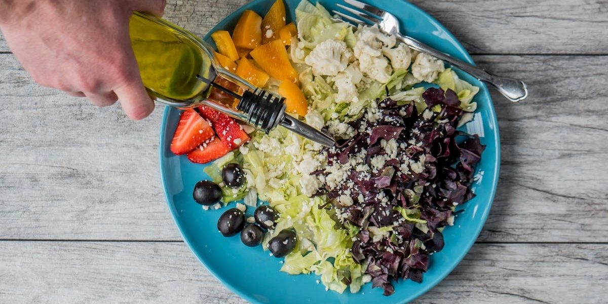 A vibrant vegetable salad with olives, feta, and strawberries being drizzled with olive oil.