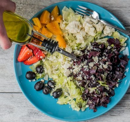 A vibrant vegetable salad with olives, feta, and strawberries being drizzled with olive oil.
